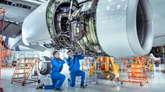 Apprentice aircraft maintenance engineers work underneath large jet engine.