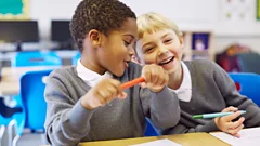Two children in primary school classroom with pens joking together