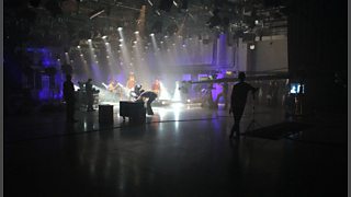 A large studio with a rock band playing. A variety of cameras and equipment are in the foreground.