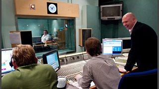Three technical staff at audio controls look on into the studio through its glass window.
