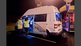 A Volkswagen Transporter van with a large satellite on top. BBC crew are working around it.