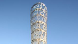 A round tubular sculpture with words encompassing it, seen against the sky.