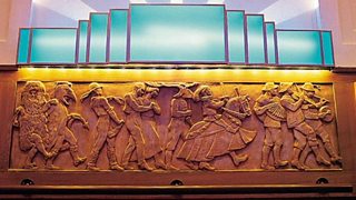 A frieze consisting of figures dancing in a procession in costumes and fancy dress. Above is an art deco light.