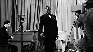 A black man, Edric Connor, sings beside a piano. A cameraman records him.