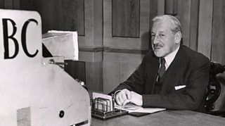 A suited politician sat at a desk talking into to a BBC television camera in a panelled room.