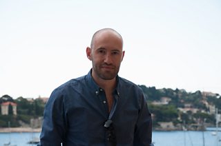 Designer, Darren Sacks looking at the camera slightly smiling and wearing a blue shirt with sea and coastline in background.