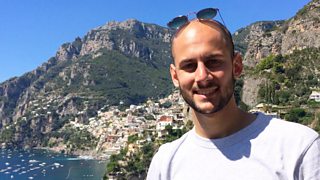 Designer, Lorenzo Strappelli looking directly at the camera smiling, wearing a grey top with sea and mountains in background.