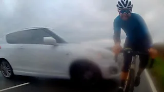 A white car slides sideways across a road, narrowly missing a cyclist who is facing the camera.