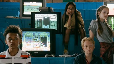 A group of children sit around 1980s TV sets showing retro video games.
