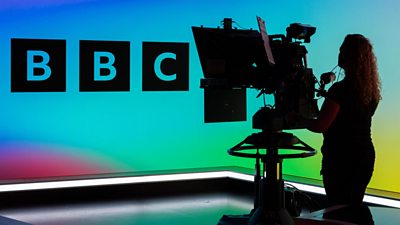Silhouette of a BBC colleague operating a camera in front of a BBC logo