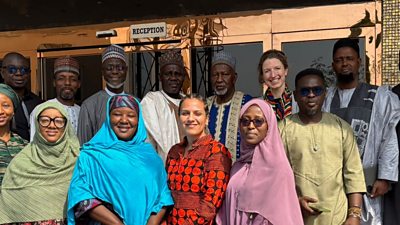 A group of men and women are smiling facing the camera, some in traditional Nigerian dress
