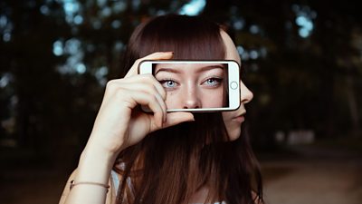 A side profile of a person as they look to the right of the image. They are holding a mobile phone alongside their head, obscuring their eyes and ear. The mobile phone is showing a close up image of the person's eyes and nose, looking straight out of the phone towards us, the person viewing this image. It creates an unsettling effect of feeling watched or viewed by the person in the picture, despite them having their head turned away from us.