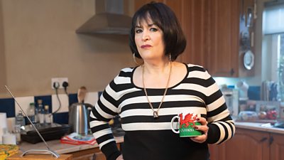 Ruth Jones stands in a kitchen as Nessa, holding a mug bearing the Welsh flag and text reading Cymru