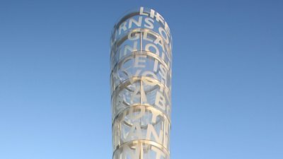A round tubular sculpture with words encompassing it, seen against the sky.