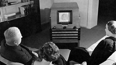 A family sit around an old black and white television in a sitting room. The television screen is tiny compared to the box and cabinet it sits in.