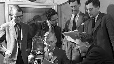 Six suited men gathered around a microphone with scripts. 