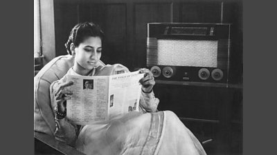 An Indian woman in a sari reads a magazine 'the voice of london' while listening to a large wireless radio. 