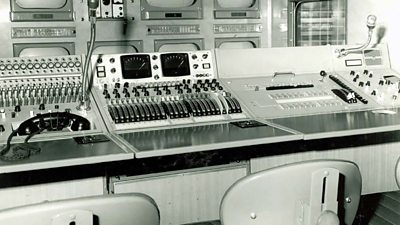 Interior of a TV outside broadcast van with a row of TV screens, various sliders and buttons on a panel and a desk with three seats.