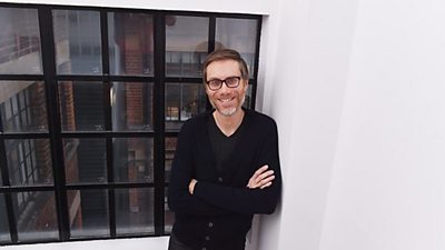  A man with a short beard and glasses wearing a black t-shirt and black cardigan standing by a window