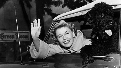 black and white image of Doris Day waving out of a car while her black poodle looks out the window with its tongue out