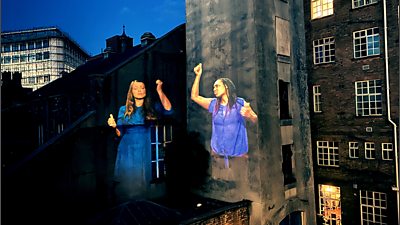 Two figures use sign language projected onto the side of a building