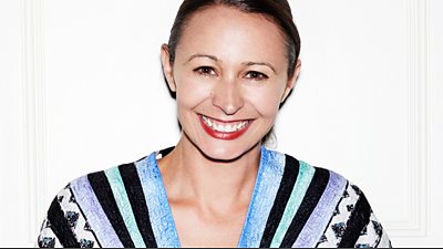 Mid shot of Caroline Rush wearing a black and blue striped top - smiling and standing in front of a white background.