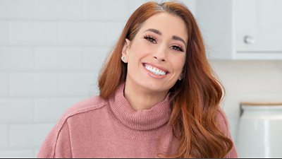 Stacey Solomon smiling wearing a pink jumper and sitting in a white kitchen