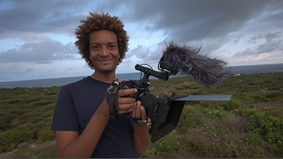 Jahawi is standing on moorland overlooking the sea, he's holding a camera with a microphone and wind muffler attached