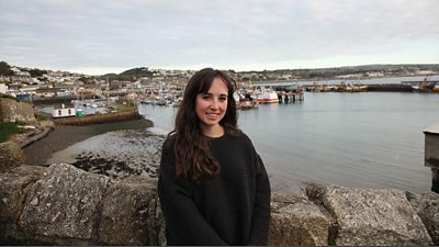 Megan has long brown hair and wears a dark jumper, she is standing in front of a small fishing harbour