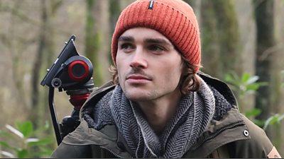 Jack is wearing an orange woolly hat, grey scarf and green coat, he is standing in front of a camera tripod