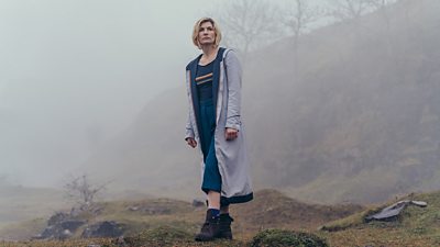 Jodie Whittaker's Thirteenth Doctor stands on a foggy landscape in her full length grey coat