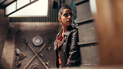 Mandip Gill's Yaz hides around a corner wearing a leather jacket and orange blouse. Spears and shields are hanging on the wall behind her.