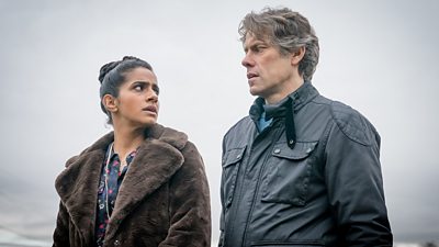 Doctor Who's Yaz and Dan stand outside in jackets. Her jacket is brown and soft while his is black and sleek. They're exchanging a worried look.