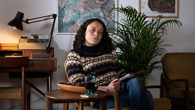 Rochenda Sandall sits in a wooden chair in what looks like an office. She's wering a black, yellow, brown and beige patterned wool jumper and is holding what looks like a digital tablet.