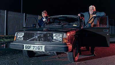 Sue Jenkins and Paul Broughton stand by a car with both the driver and passenger doors open. They're staring at something that appears to be shining a bright light on their black car.