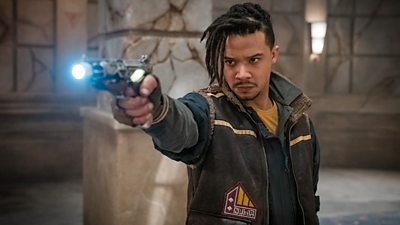 Jacob Anderson's Vinder points a gun with a glowing barrel at an unseen target. He's wearing a military style waistcoat over a jacket and his hair hangs in short dreadlocks.