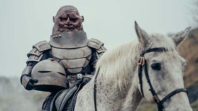 A Sontaran sits on a white horse wearing armour and carrying a helmet under their arm.