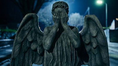 A stone weeping angel stands in the middle of a road with its hands over its eyes.