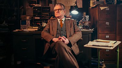 Kevin McNally reclines in a chair in a study in Doctor Who Flux. He's wearing a brown suit and is surrounded by newspapers and books.