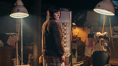 Annabel Scholey stands in front of a control panel. Two large lights beam down upon her and a chair beside her, which appears to be connected to wires and features some straps.