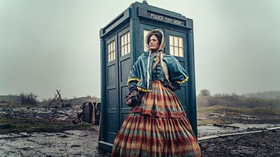 A woman in a large multicoloured hooped skirted dress, a bonnet and blue shawl  stands outside the TARDIS on a barren landscape.