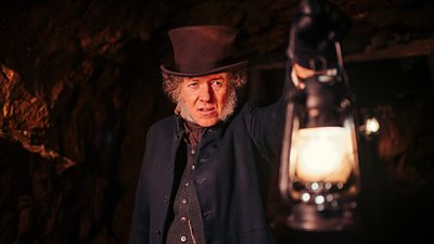 A man in a brown top hat and navy jacket holds a lamp up in a dark cave.