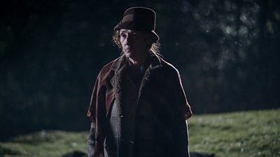 A woman in a coat, hat and shawl stands half in shadow in a field or park on a dark night.