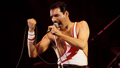 Queen singer Freddie Mercury clutches a microphone cable in his hand as he sings on stage in a white tank top with a red lightning bolt on it.