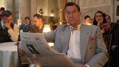 Dominic West sits at a small table with a white tablecloth reading a newspaper.