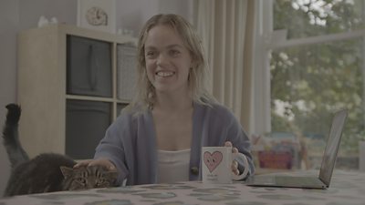 Paralympian Ellie Simmonds sits at a table with a mug in hand, stroking a cat who is sneaking a glance over the top of the table.