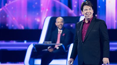 Michael McIntyre presents gameshow The Wheel, wearing a dark suit jacket and a maroon shirt.