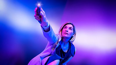 Jodie Whittaker's Doctor is bathed in blue and purple light, holding her sonic screwdriver aloft and ready for action