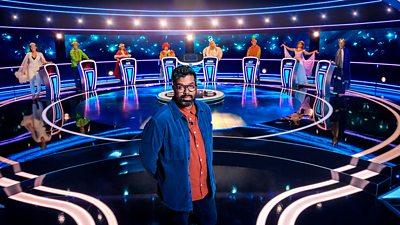 Host Romesh Ranganthan stands on the set of The Weakest Link where contestants stand at podiums in Christmas fancy dress