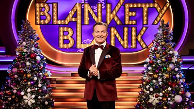 Blankety Blank host Bradley Walsh wears a red velvet jacket and stands between two decorated Christmas trees on the set of the gameshow